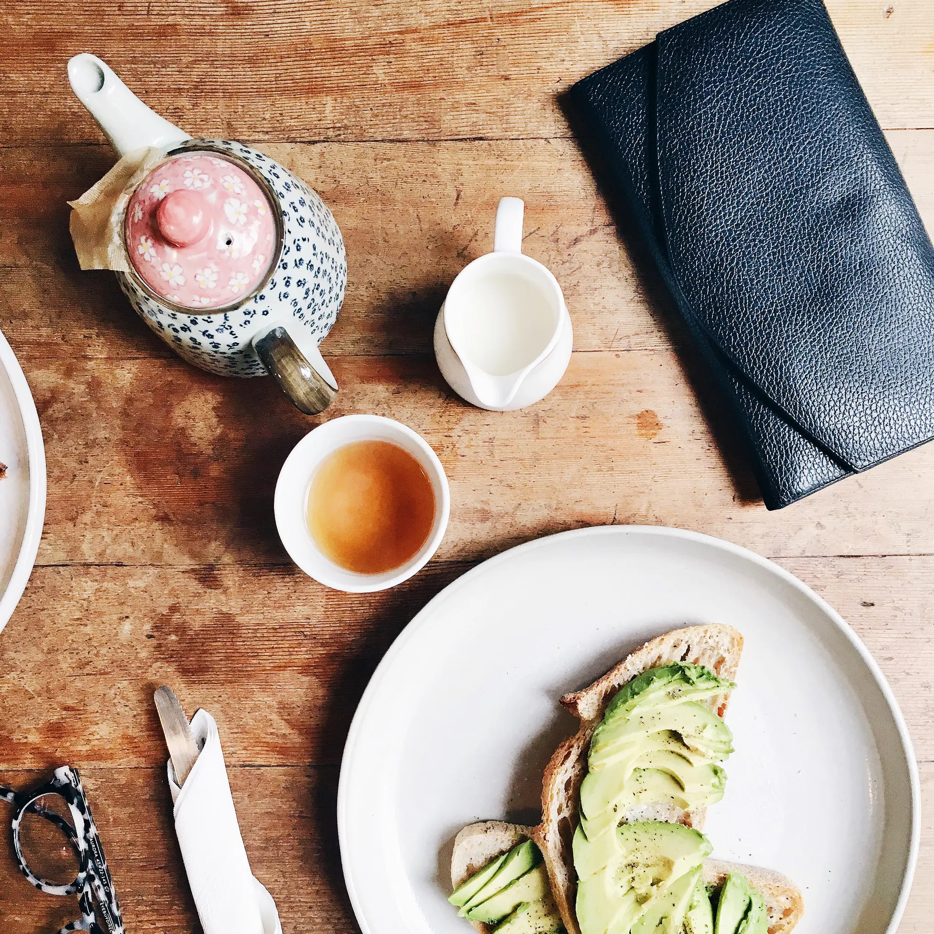 Une théière fleurie, un pot de lait, une tasse de thé et une tartine d’avocat sur la table de bois
